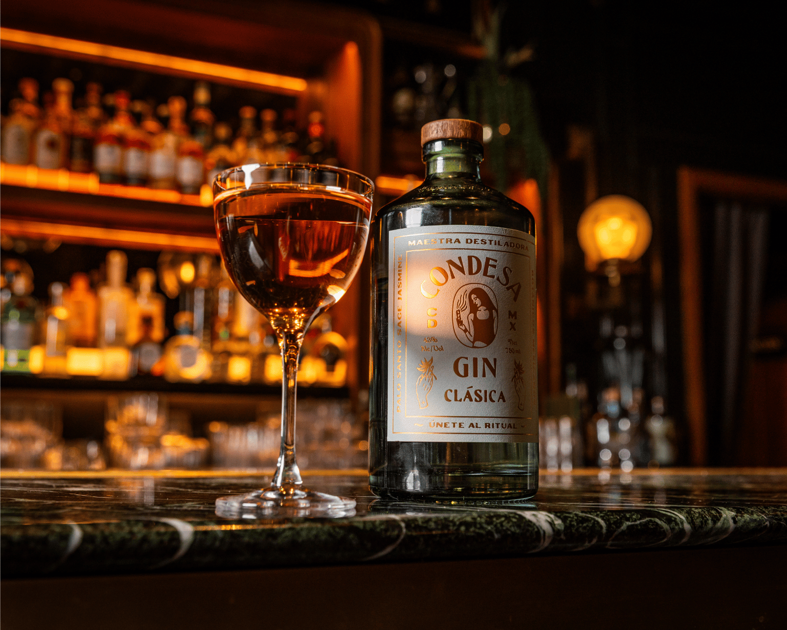 Chicago 13 A bottle of Condesa Gin Clásica with a wooden cap stands on a marble bar counter next to a filled stemmed cocktail glass, with a softly lit, out-of-focus bar and liquor shelves glowing warm orange in the background.