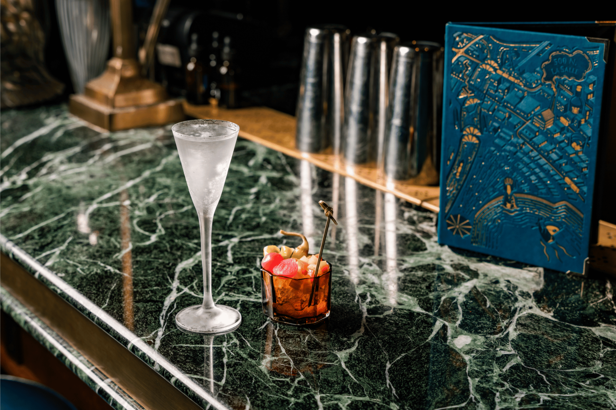 A chilled martini glass and a short amber tumbler filled with colorful garnishes sit on a green marble bar top. In the background, a vibrant blue cocktail menu embossed with a map of 500 N. 
