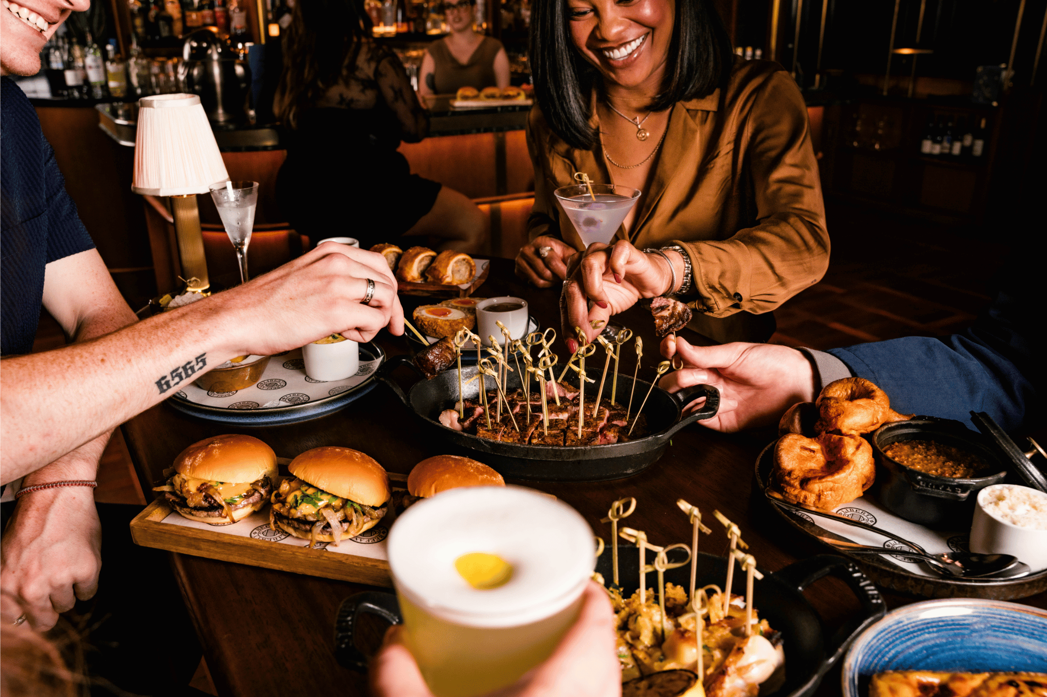 A lively group of people are enjoying cocktails and a variety of shareable bar bites at a table. The spread includes sliders, Yorkshire puddings, and skewers served in a cast iron pan.