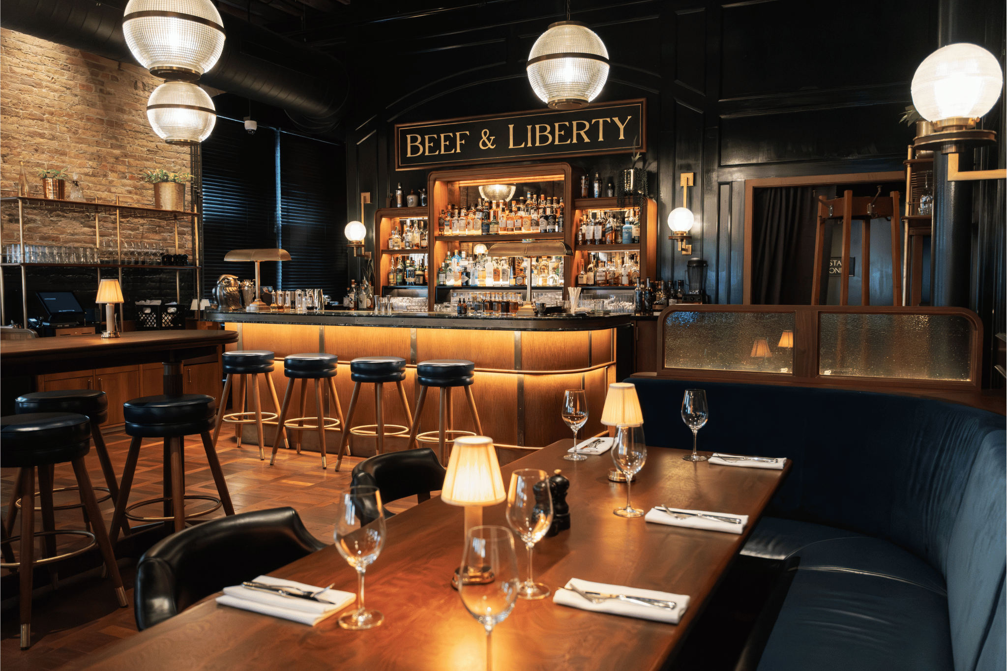 An interior shot featuring a warmly lit bar with shelves of spirits, high wooden stools, and booth seating area set with glasses and napkins. The space features exposed brick, vintage lighting fixtures and dark paneling.