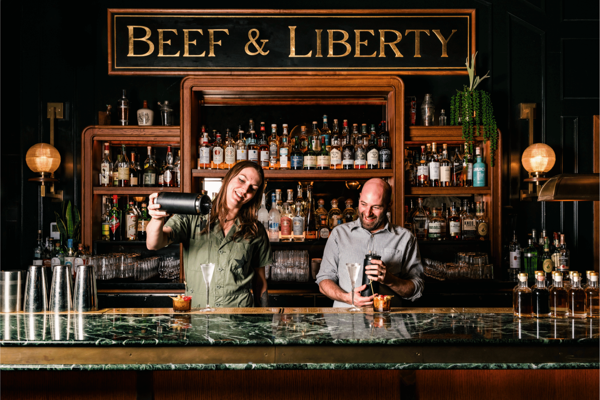 Two bartenders stand behind a green marble bar, mixing drinks in front of shelves filled with bottles of spirits. Above them, a large gold sign reads "Beef & Liberty".