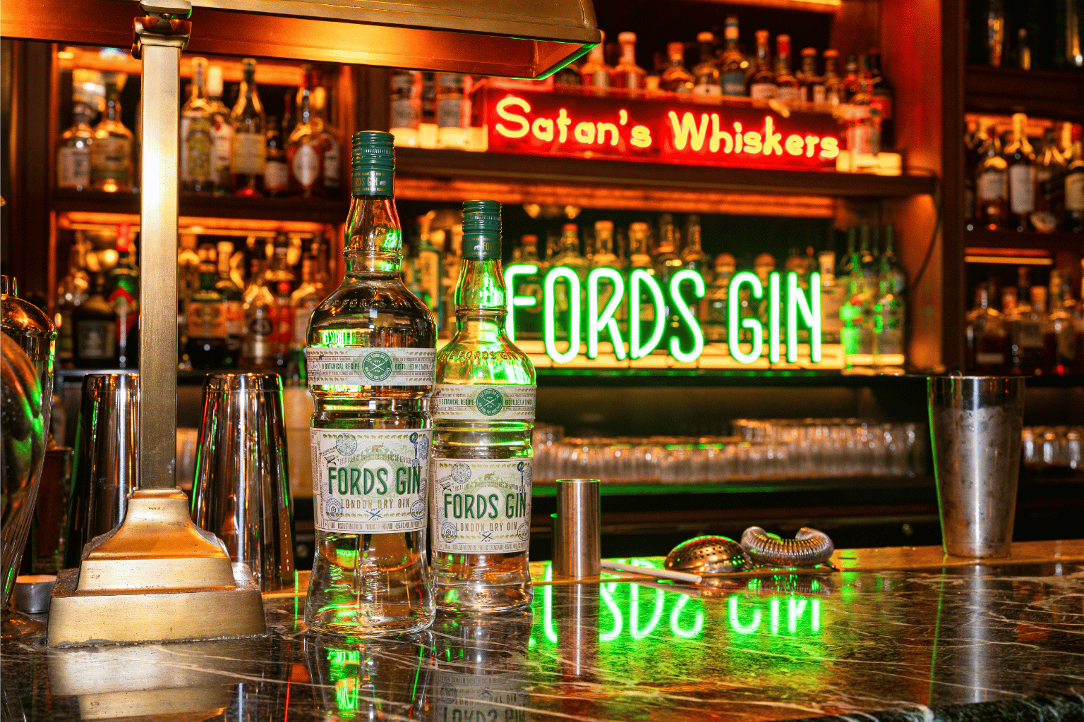 Two bottles of Fords Gin are displayed on a marble bar next to cocktail tools and shakers, with shelves of liquor in the background. In the background neon signs reading "Satan's Whiskers" and "FORDS GIN". 